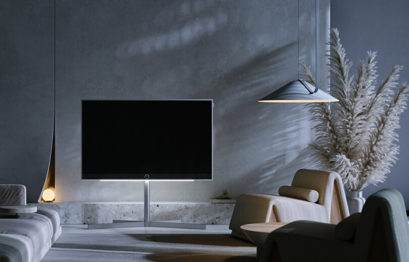 Minimalist living room with a contemporary design featuring a Loewe Stellar OLED television on a stand, modern furniture, a pendant lamp, and decorative pampas grass against a gray wall.
