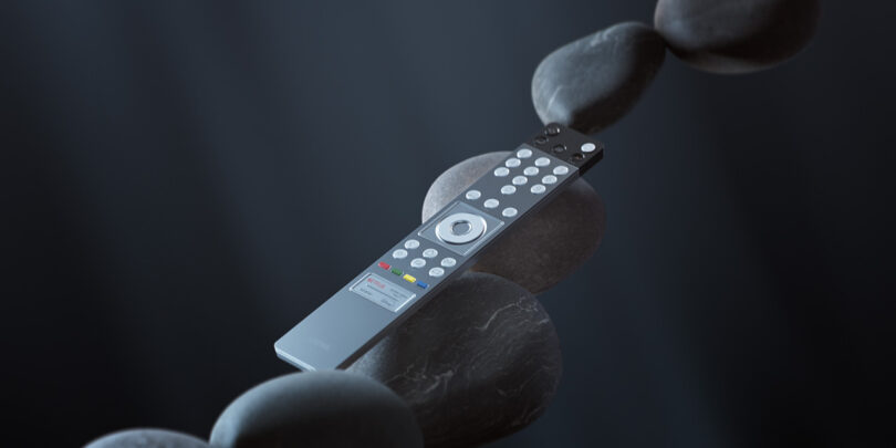 A Loewe Stellar OLED television remote control rests on a stack of smooth, black stones in a balanced arrangement.