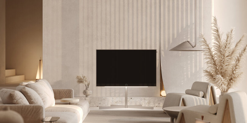A modern living room featuring a Loewe Stellar OLED television, a white sofa, two armchairs, and contemporary decor elements, including pampas grass and a hanging light fixture.