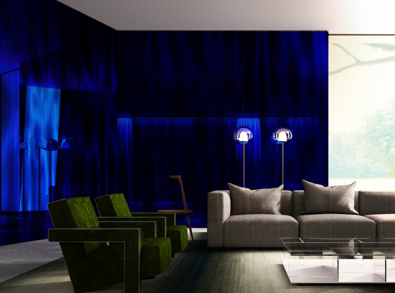 A modern living room with deep blue walls, green armchairs, a gray sofa, a glass coffee table, and two standing lamps near a large window.