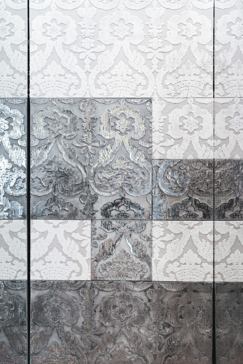 Close-up of a patterned wall featuring square tiles with intricate floral and geometric designs in varying shades of gray and white. The texture of the tiles appears metallic and reflective.