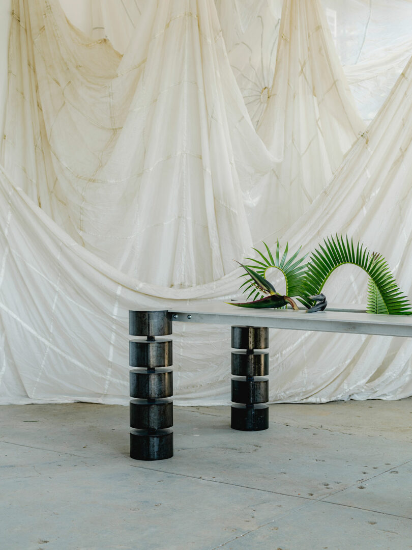 A minimalist black table with cylindrical legs stands on a concrete floor, adorned with green palm leaves; sheer white curtains hang in the background.