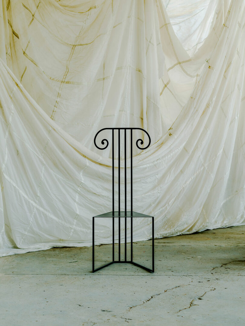 A minimalist black metal chair with decorative curls at the top sits in front of draped white fabric in an indoor setting with a polished concrete floor.