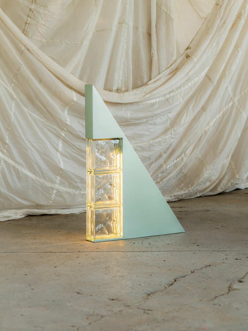 A geometric sculpture featuring a tall, triangular panel with three rectangular glass blocks at the base, set against a draped white fabric background.