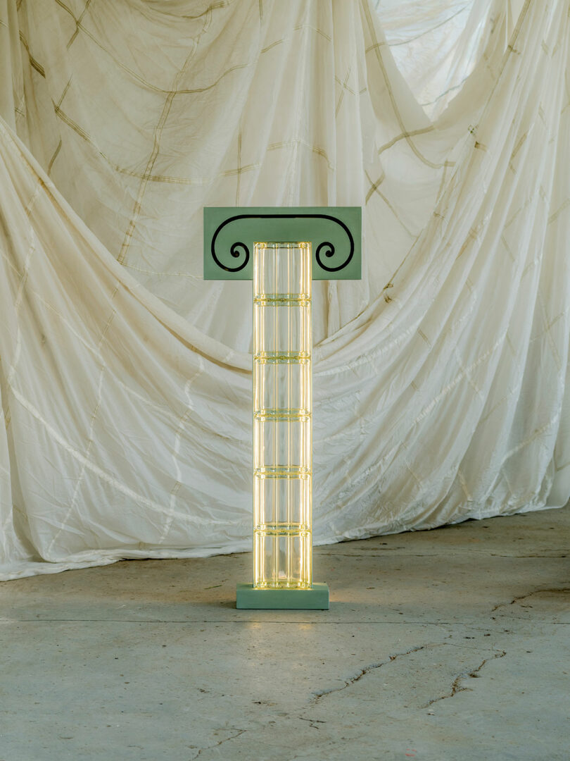 A decorative column-shaped light fixture with green accents and an ornate top, standing in front of a backdrop made of draped white fabric.