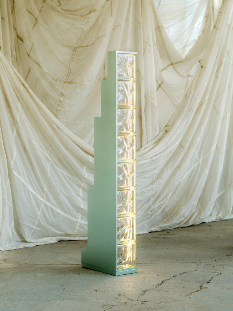 A tall, rectangular art installation featuring stacked acrylic boxes with intricate light patterns set against a draped fabric backdrop.