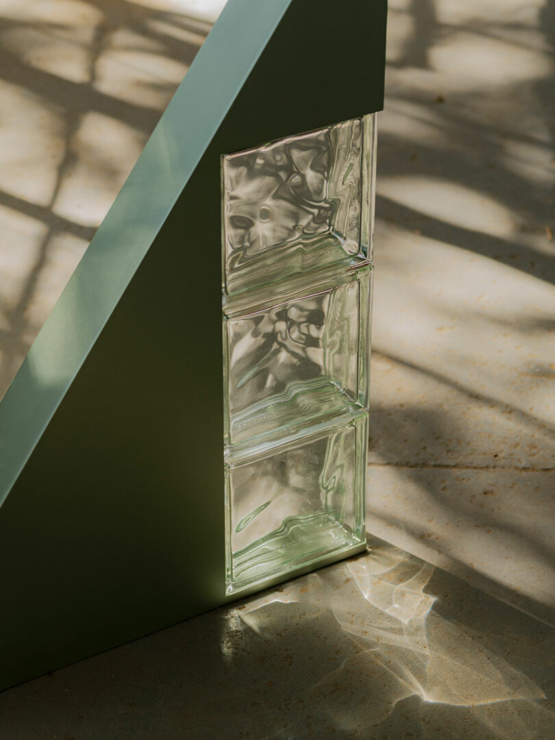 A green metal structure with three adjacent glass blocks casting shadows on a sunlit floor.
