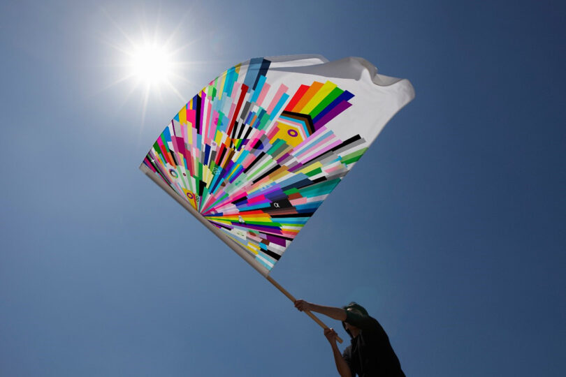 Person holding a flag with a vibrant, geometric pattern against a clear blue sky and bright sun.
