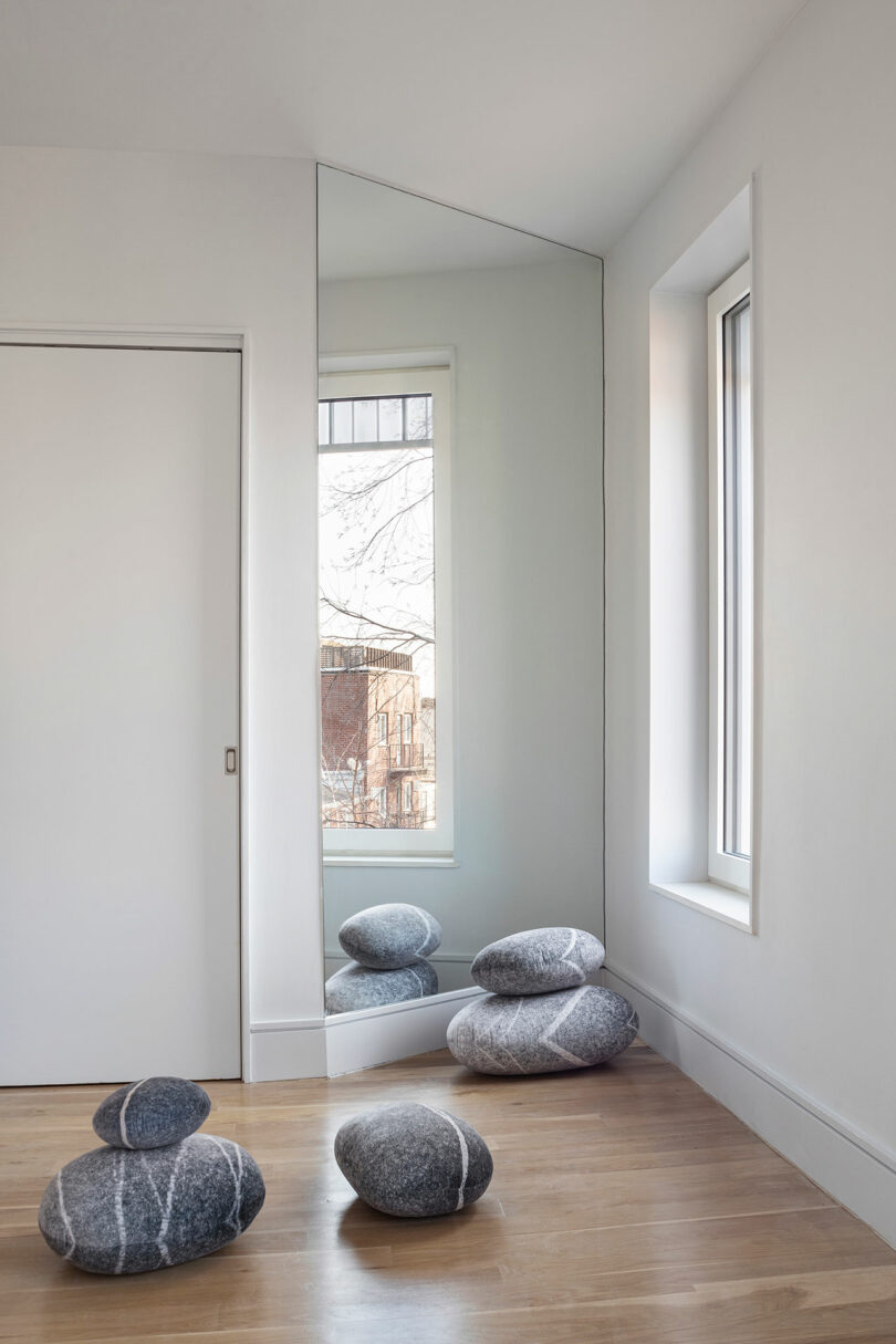 A minimalist room with a large wall mirror reflecting a window and five gray stone-like cushions on a wooden floor. The room features white walls and a sliding door.
