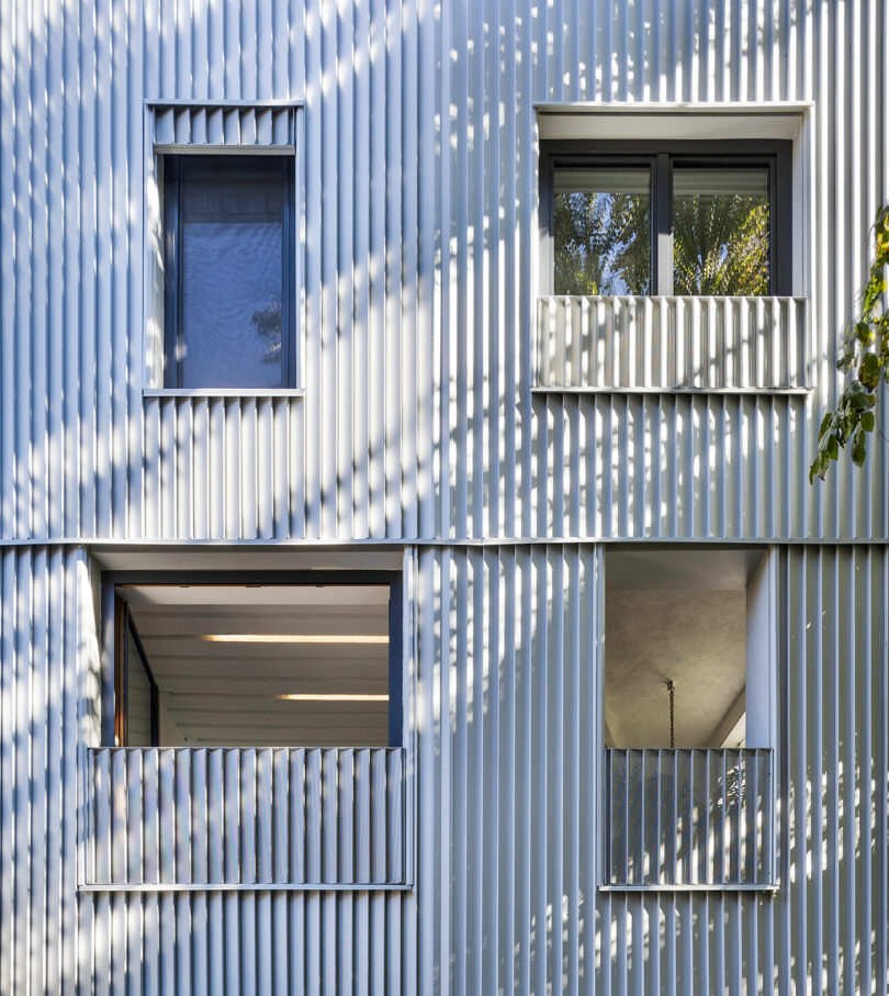 The image shows a modern building facade featuring a corrugated metal design with several windows and balconies, partially shaded by tree leaves.