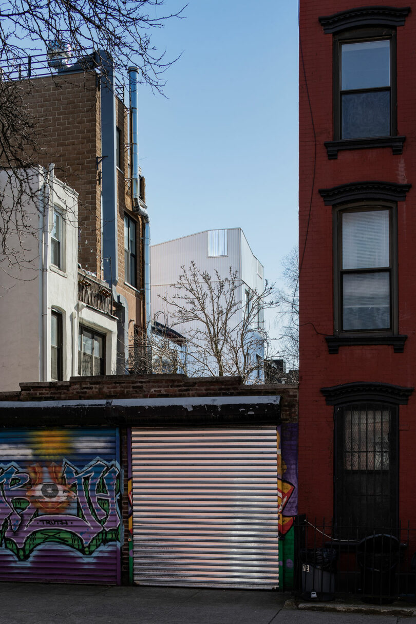 A narrow alleyway featuring a closed metal roll-up door with graffiti, flanked by multistory buildings with exposed pipes and a red brick facade. The background includes modern architecture and bare trees.