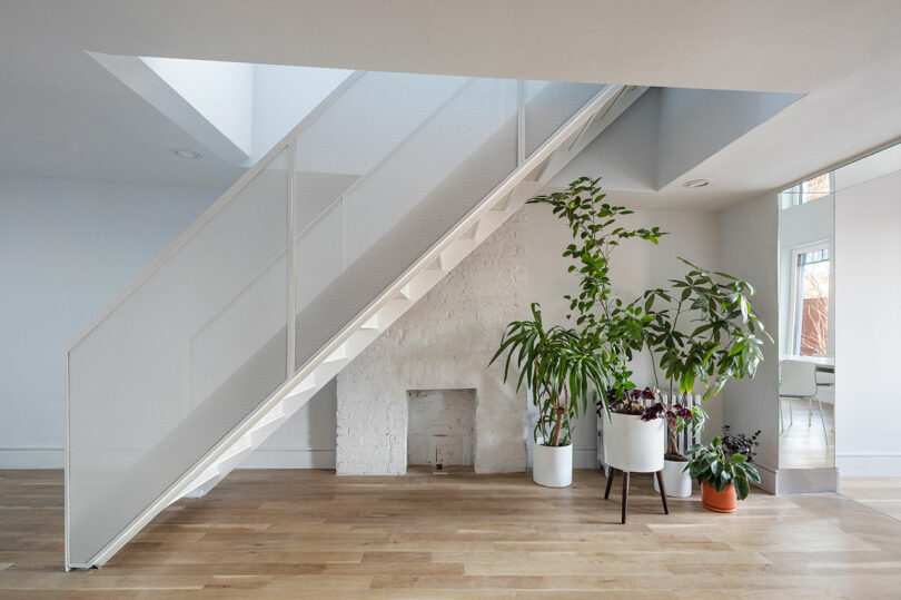 A minimalist staircase with a white railing and steps ascends next to a white brick wall. Several potted plants are arranged below the staircase on a light wood floor.