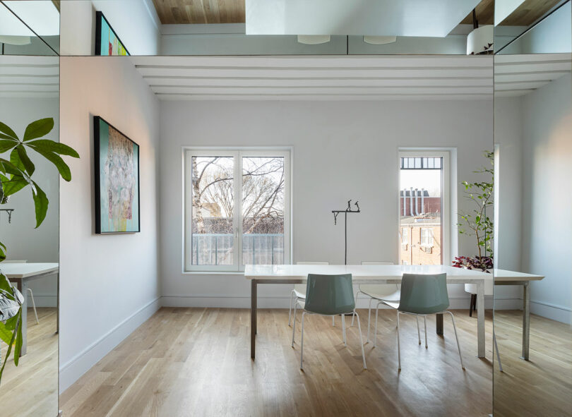 Modern, minimalistic room with a white table, two blue chairs, wall art, and a large mirror reflecting the space. Natural light enters through two windows, and there are hints of greenery.
