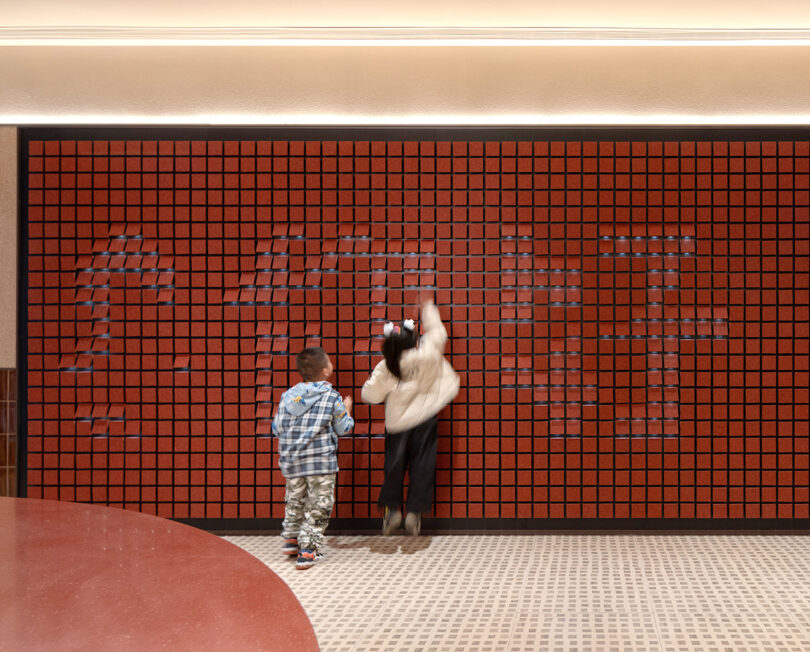 Two children interact with a red pixelated wall display at the NI HAO hotel. The girl reaches up to touch the tiles while the boy stands beside her watching. The floor is partially red and partially checkered.