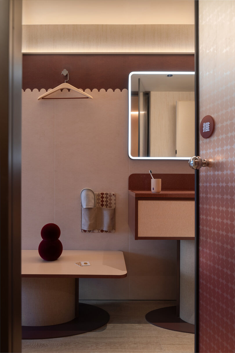 Modern bathroom at the NI HAO hotel with a beige and brown color scheme. A wall-mounted sink with a lit mirror, towel holder, and a small table with two decorative balls are visible. The door is ajar.