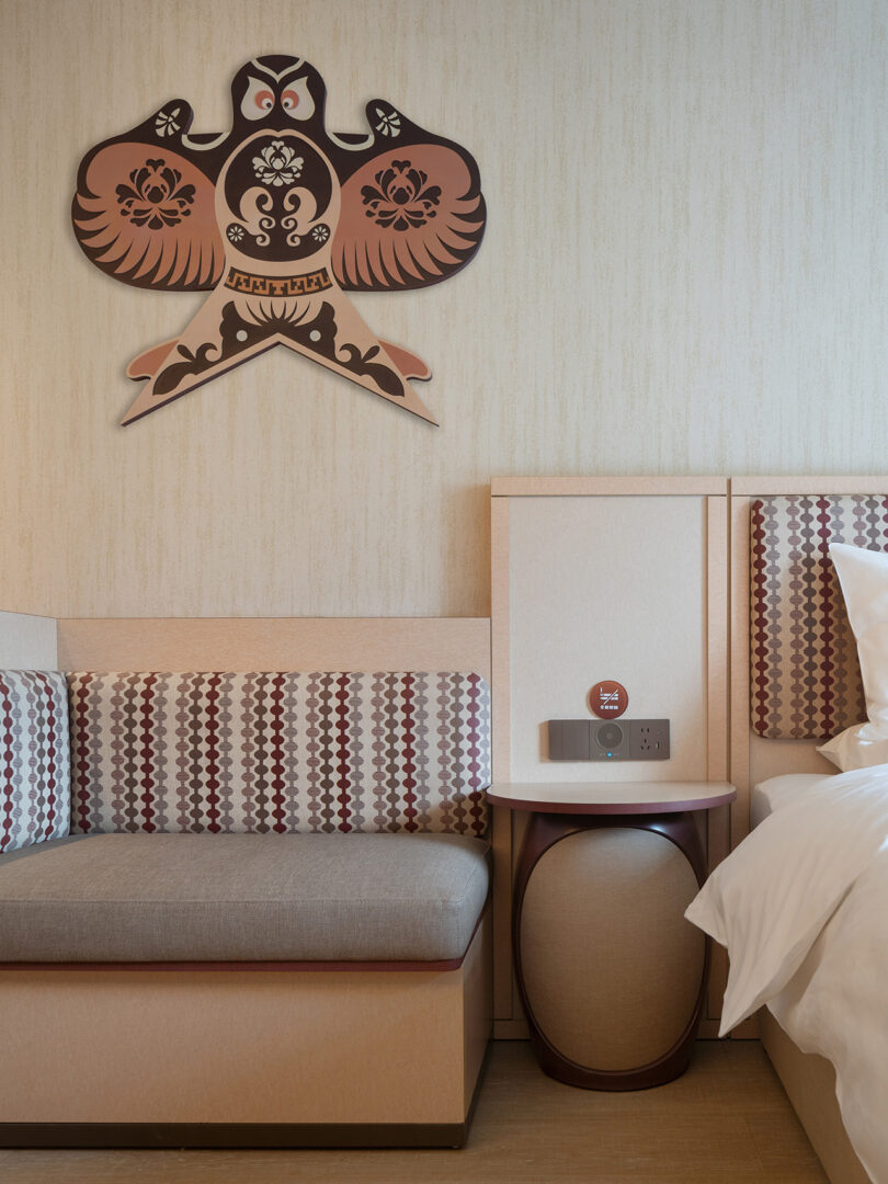 A cozy corner at NI HAO hotel featuring a cushioned bench next to a bedside table with a circular base, all beneath an elegant wall art piece of a stylized bird.
