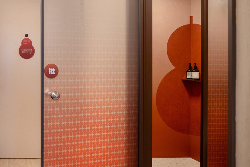 A door with a geometric pattern opens to a modern room with red walls and a shelf holding two bottles, echoing the elegant design of the NI HAO hotel.