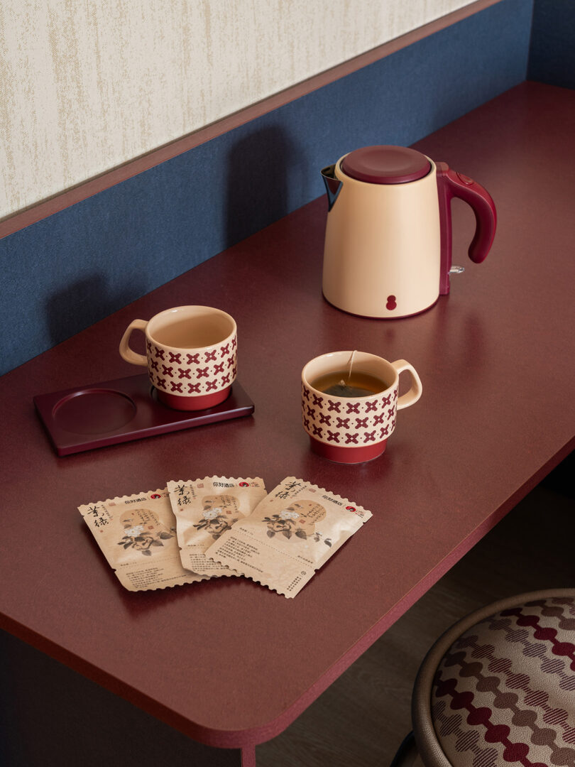 A red desk with two patterned mugs, tea bags, a red and beige electric kettle, and a cup coaster creates a cozy corner reminiscent of the welcoming ambiance at NI HAO hotel.
