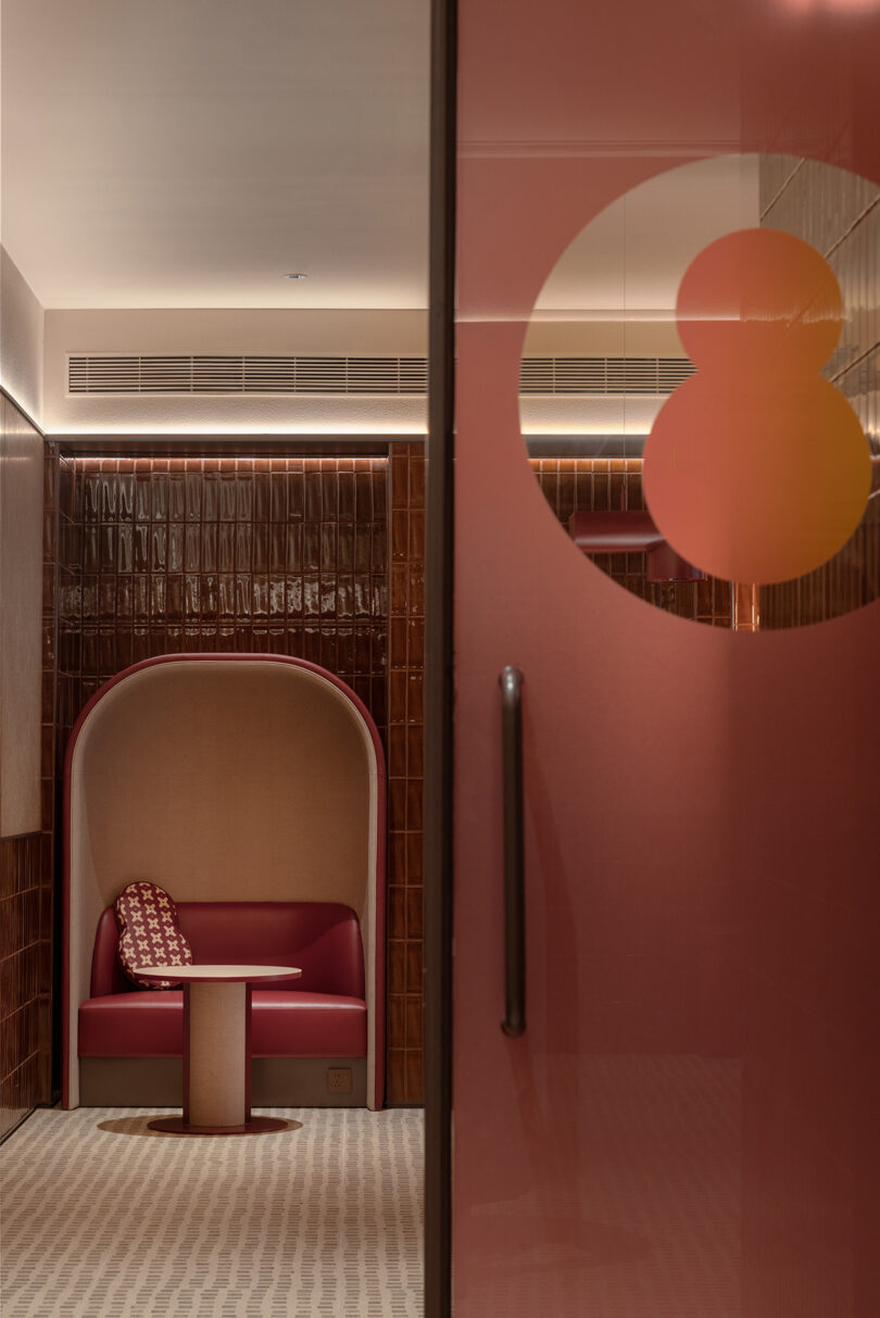 Modern seating nook with red leather booth and small table, set against a textured brown tile wall, seen through a glass door with the number eight at NI HAO hotel.