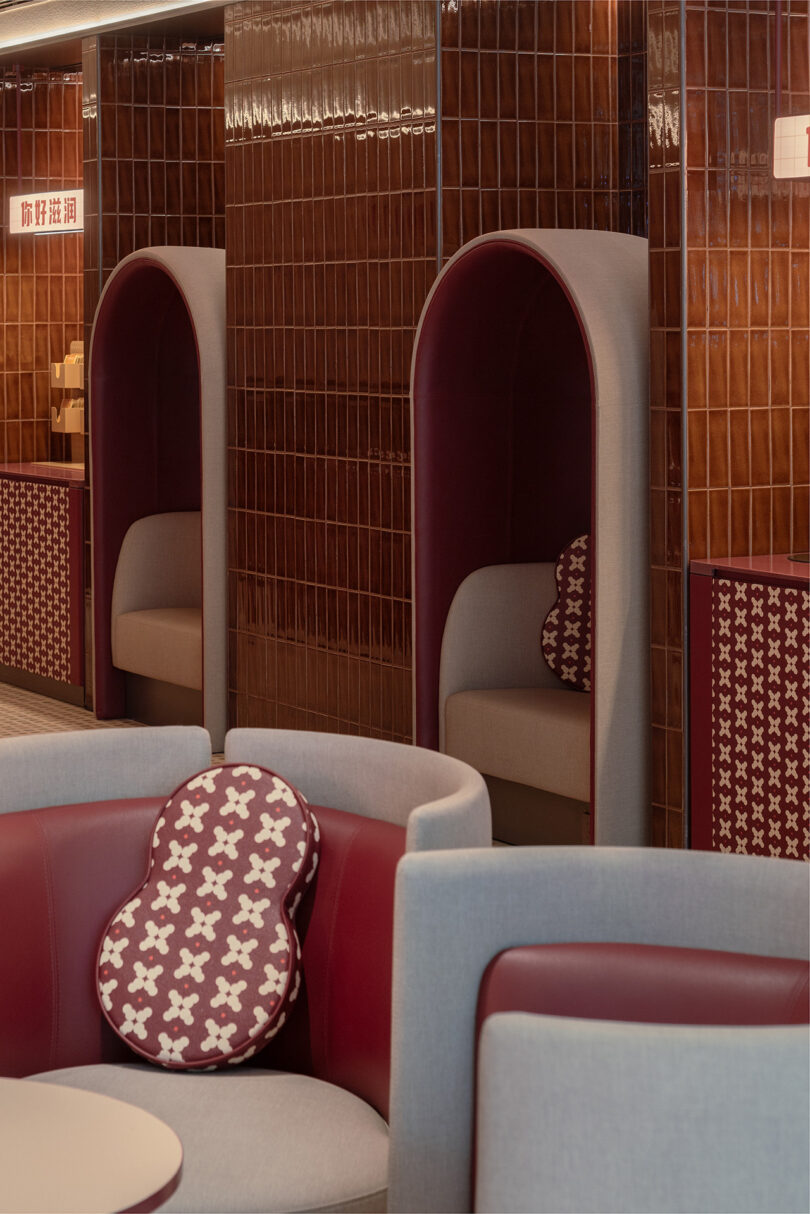 The image shows seating areas with maroon and grey upholstered chairs, some enclosed by curved panels, and patterned cushions. The background features tiled walls and a sign with Chinese characters, suggesting this might be the cozy lounge at NI HAO hotel.