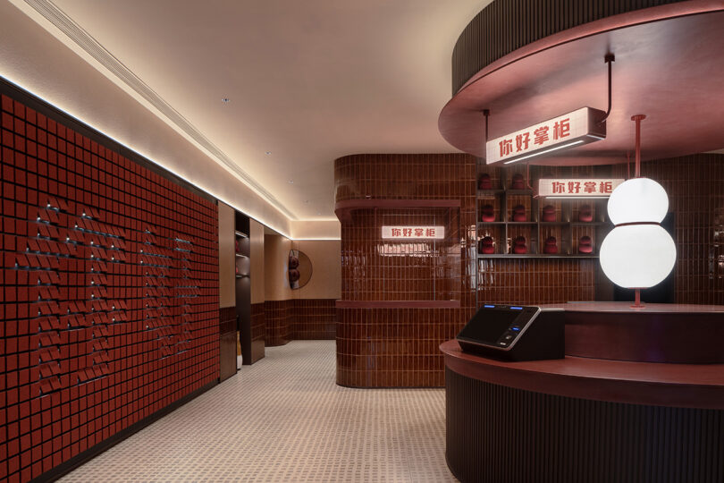 A modern interior at the NI HAO hotel features red tile walls, a curved reception desk, a digital kiosk, and spherical pendant lights. Chinese characters are elegantly displayed on illuminated signs in the background.