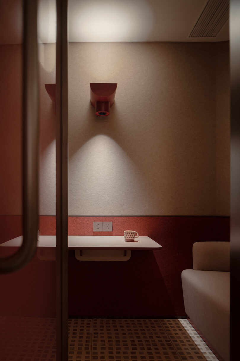 A small, modern room at the NI HAO hotel boasts a red and beige color scheme, featuring a wall-mounted desk, a single light, a power outlet, a mug, and part of a grey couch.