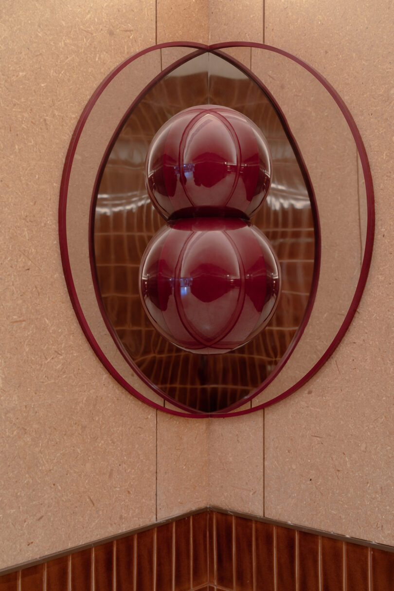 A modern wall-mounted art piece featuring two stacked, glossy burgundy spheres encased in a reddish circular frame, displayed on a textured beige wall above brown tiles at the NI HAO hotel.