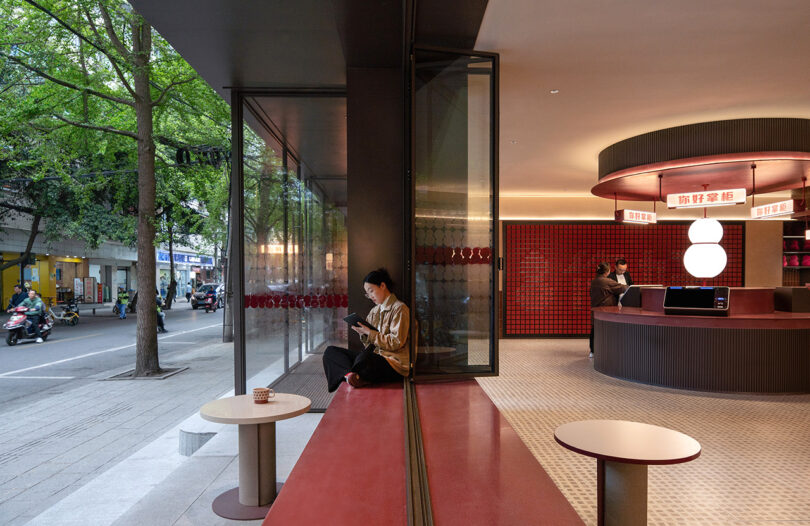 A person is sitting inside a modern café at the NI HAO hotel, reading a book. The café features red seating and a stylish round counter. The window offers a view of the street outside, with trees and parked scooters.