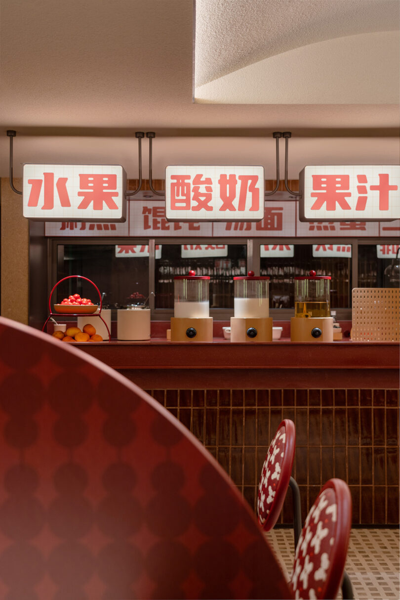Interior of a cafe with red and white decor, featuring a counter with fruit, drinks, and snack items. Signs in Chinese characters hang above the counter, indicating fruit, yogurt, and juice. The cozy ambiance feels like an inviting extension of the NI HAO hotel nearby.