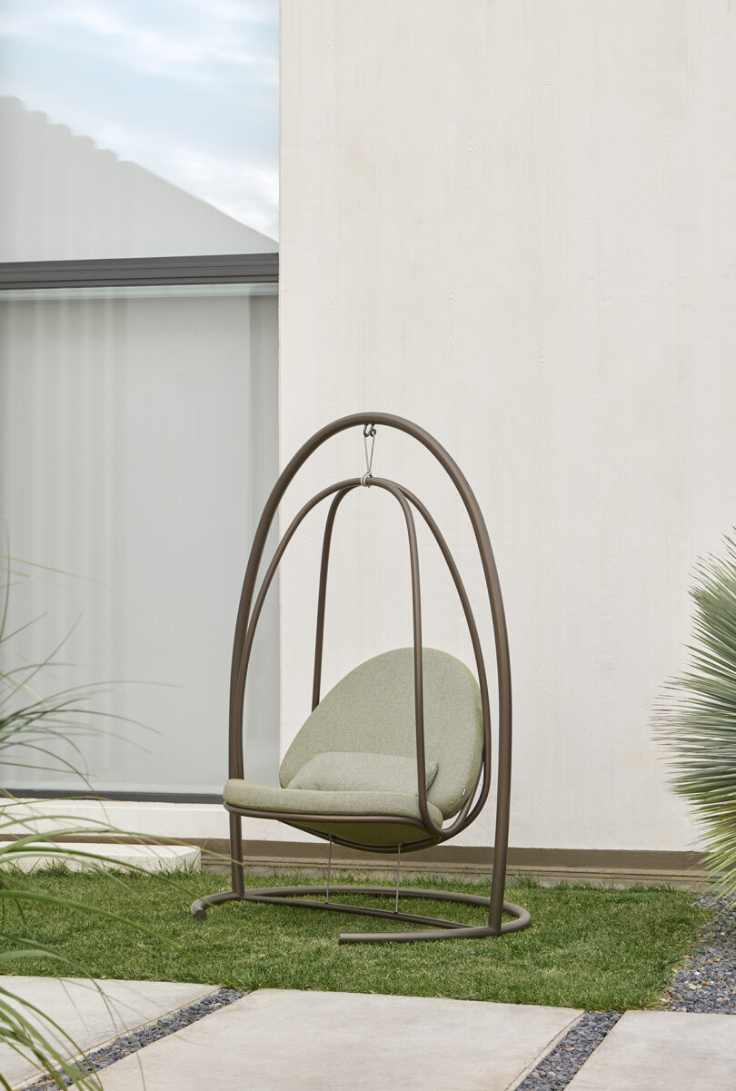 A cushioned swing chair with a metal frame is placed on a grassy area next to a concrete walkway and a light-colored exterior wall.