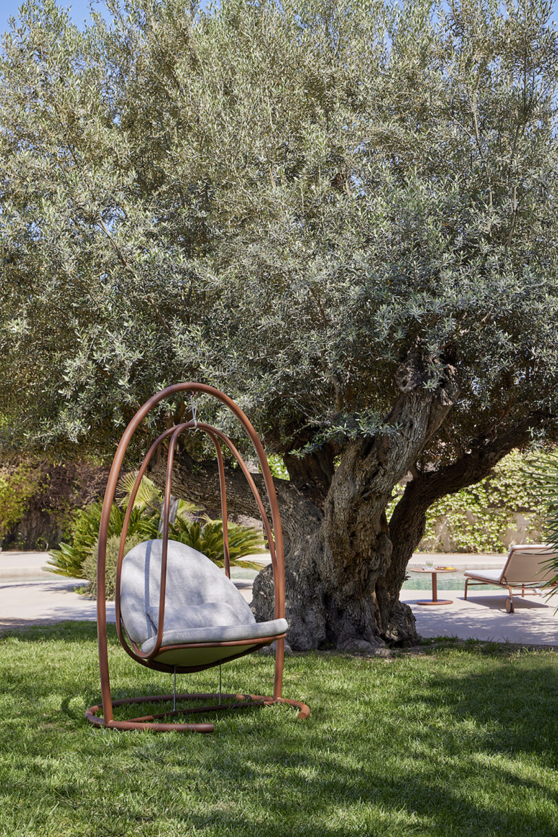 A cushioned hanging chair is placed on a lawn beneath a large tree. Another chair and a table are seen in the background beside a pool.