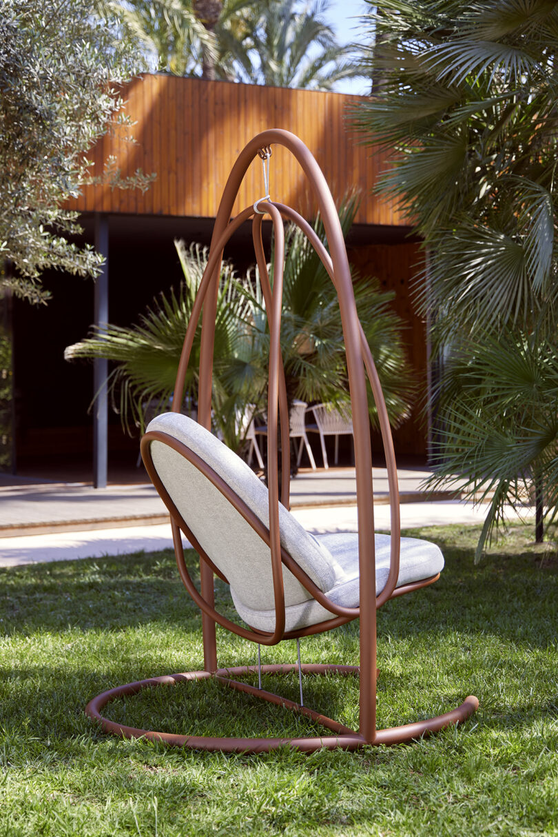 A hanging chair with a cushioned seat is suspended within a metal frame on a grassy lawn. There is a wooden building and tall green plants in the background.