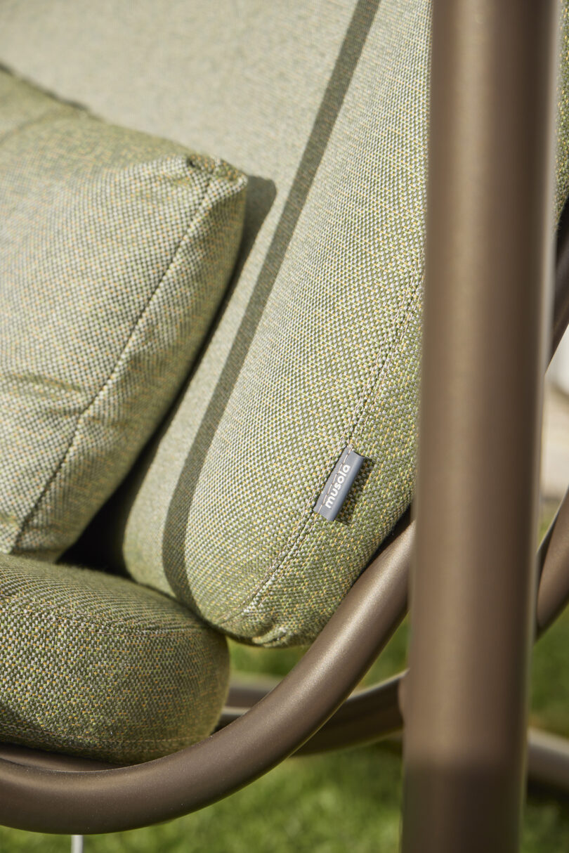 Close-up of a green cushioned outdoor chair with a textured fabric. The leg and supporting frame are brown metal, and a branded fabric tag is visible on the side of the cushion.