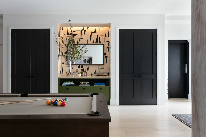 A modern room features a pool table with a set of balls and cue stick, flanked by two closed black doors and a decorated niche with a mirror, greenery, and abstract wallpaper.