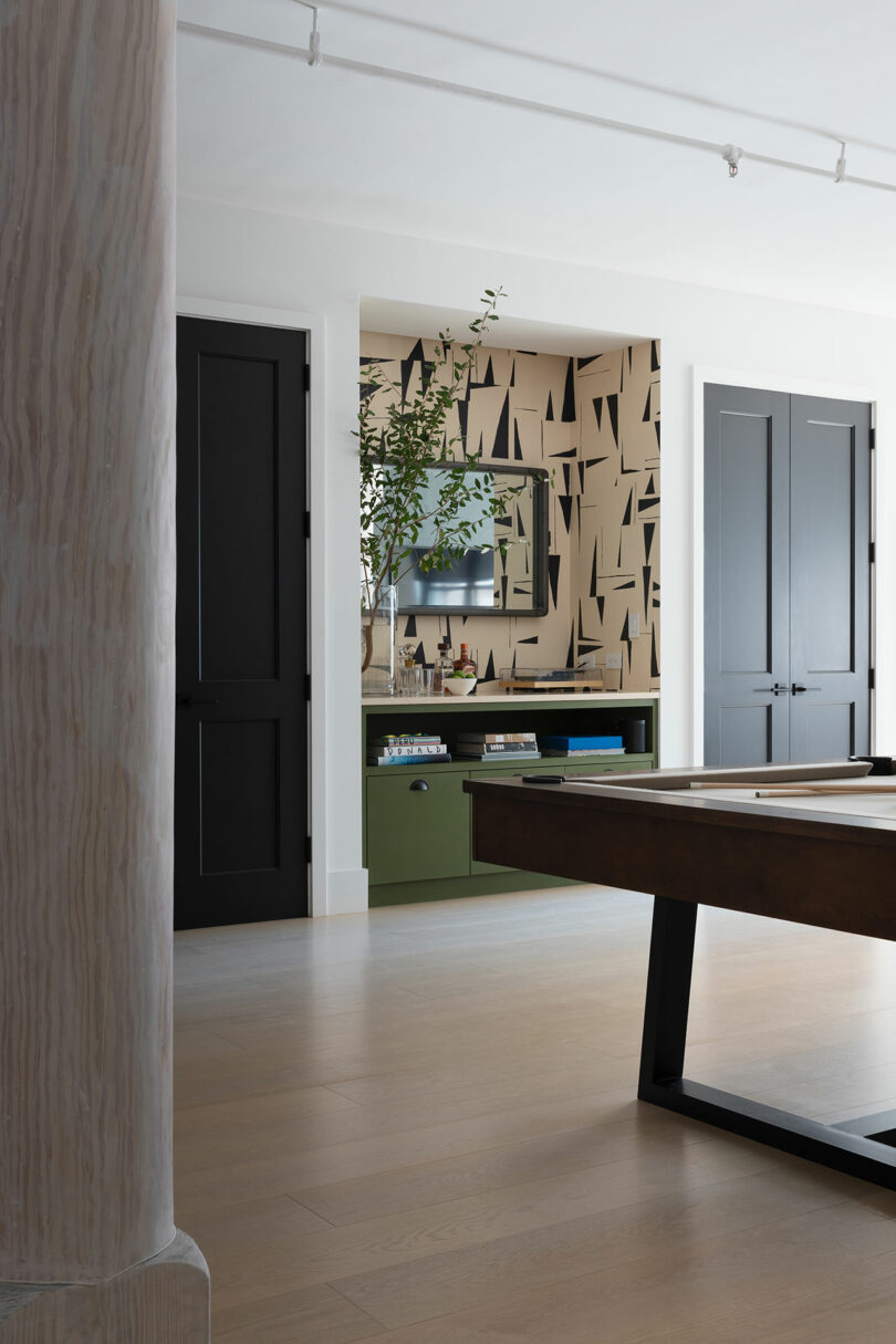A modern room featuring a pool table, black cabinets, a green console with books, a TV on a geometric-patterned wall, light hardwood floors, and track lighting on the ceiling.
