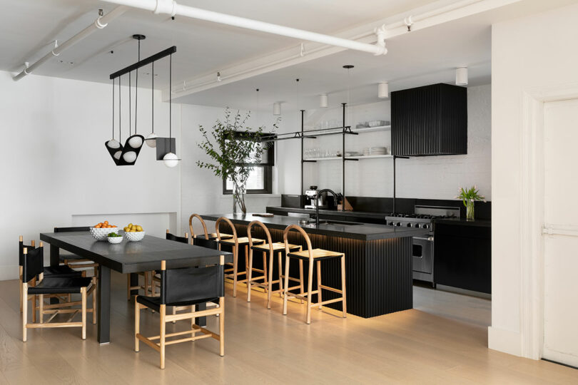 Modern kitchen and dining area with black cabinetry, light wood floors, a long black dining table with chairs, a kitchen island with stools, hanging lights, and a potted plant on the counter.