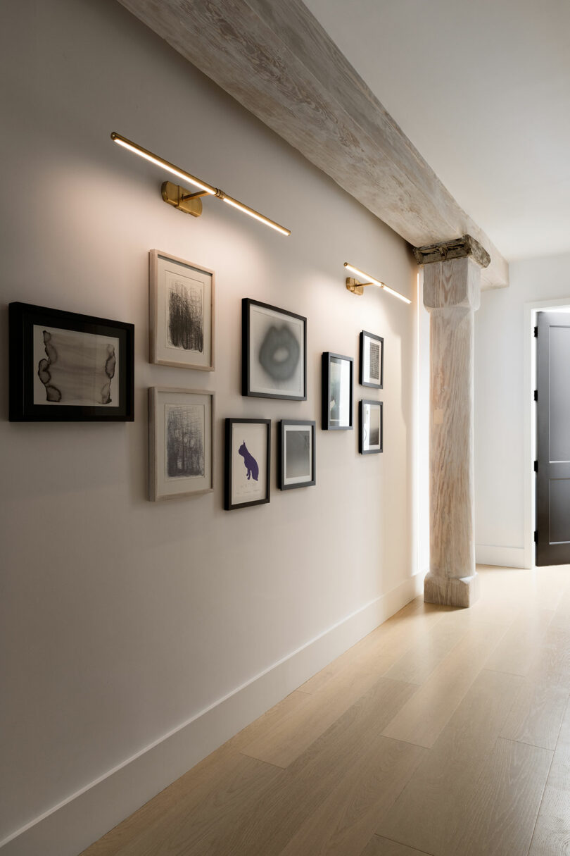 Hallway with framed artwork on the wall, illuminated by two linear lights. The space features light wood flooring and an exposed wooden beam near an open doorway.