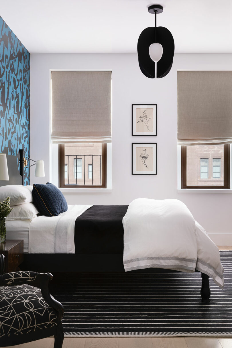 Modern bedroom with a single bed, black and white bedding, patterned accent wall, two windows with beige shades, three framed artworks, black chandelier, and a striped rug.