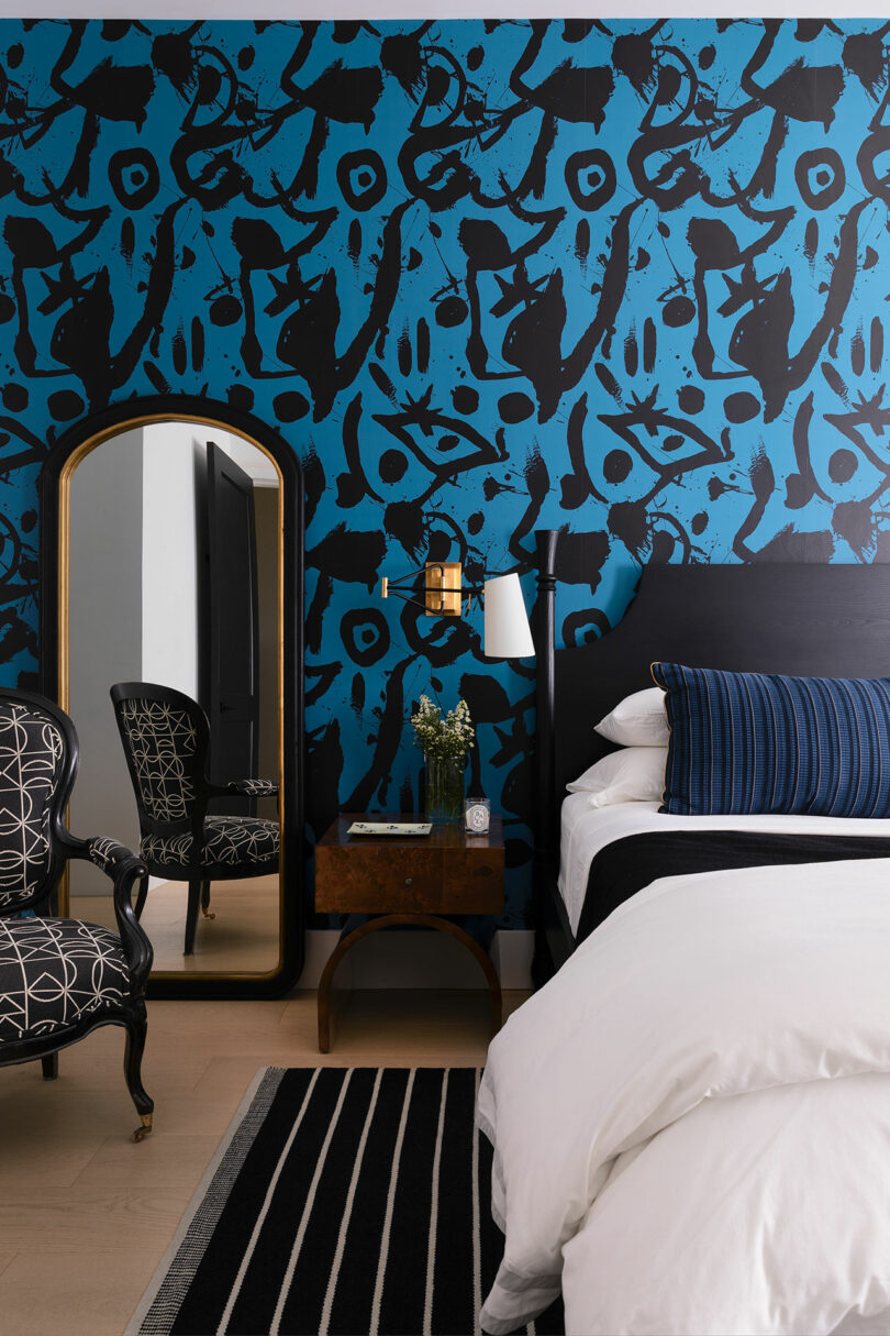 Modern bedroom with black and blue art wallpaper, a bed with white linens, a wooden nightstand with flowers, a black and white striped rug, and an ornate black chair next to a tall, gold-framed mirror.