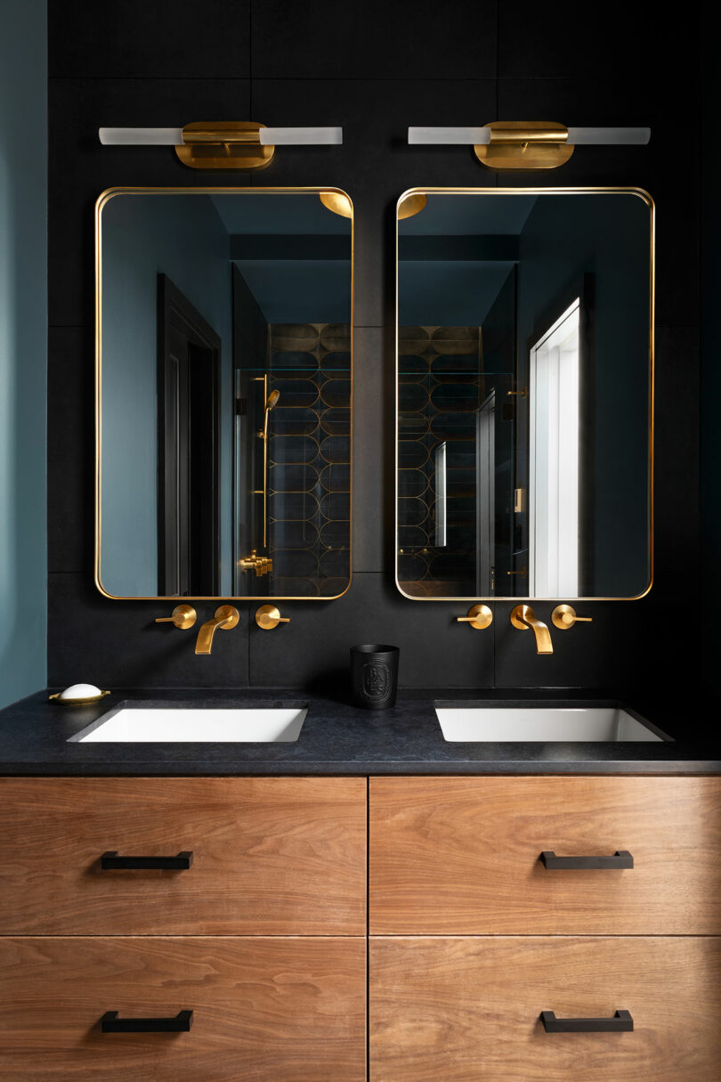 Modern bathroom with two rectangular mirrors, gold fixtures, and a wooden double-sink vanity. Light fixtures are mounted above each mirror. Black countertops contrast with the dark wall behind.