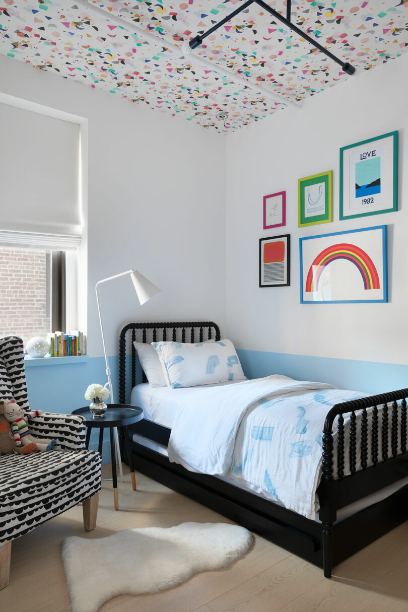 A child's bedroom with a black bed, patterned chair, white floor lamp, colorful ceiling and art, white walls, and a window with a shade. The bed has white and blue bedding with animal prints.