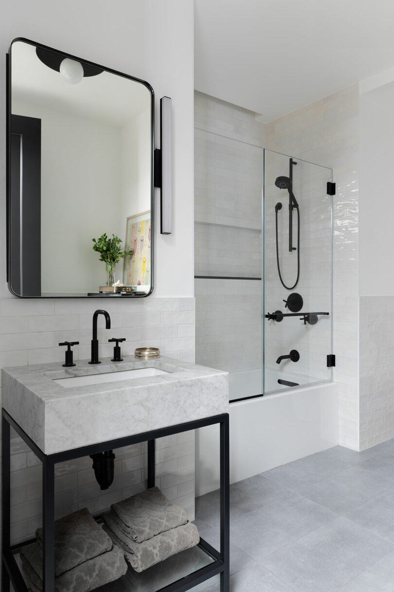 Modern bathroom with a marble-topped vanity and black fixtures, a floating shelf with towels underneath, a glass-enclosed bathtub with black shower fixtures, and a large mirror above the sink.