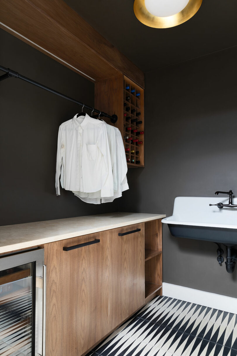 A laundry room with a dark wall, wooden cabinets, a countertop, a white sink, a hanging rail with white shirts, a built-in wine rack, and patterned floor tiles.