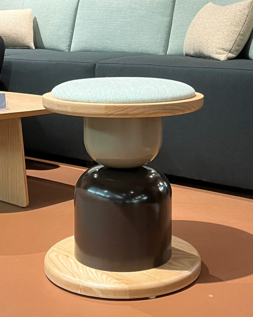 A modern stool featuring a light wooden base, a black middle section, and a cushioned seat is elegantly placed on a brown floor in front of a sofa.