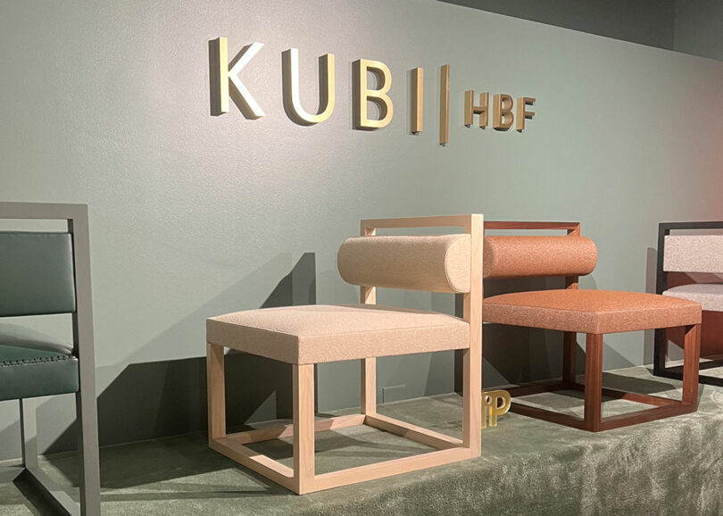 A display of modern chairs is set against a green wall with the words "KUBI HBF" prominently displayed above them, reminiscent of the stylish showcases at NeoCon. The chairs feature geometric designs and various fabric colors.