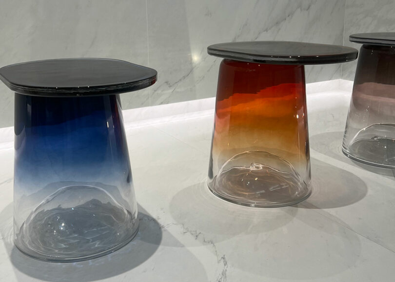 Three gradient-colored glass tables with dark round seats are displayed on a marble floor. Colors transition from blue to transparent on the left, orange to clear in the center, and purple to clear on the right.