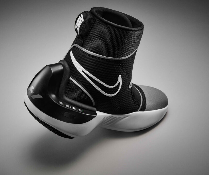 Black and white athletic boot with a high ankle collar and a large white logo on the side, featuring Nike x Hyperice recovery technology, set against a gradient grey background.