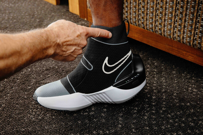 A hand points at a black and grey high-top sneaker with a white Nike logo, featuring a cushioned white sole and black heel, worn by a person with their foot resting on the floor. This exclusive Nike x Hyperice collaboration integrates advanced recovery technology for ultimate comfort.