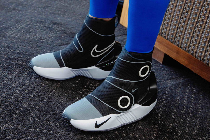 Person wearing blue leggings and black athletic shoes with white soles and Nike branding, featuring straps and a futuristic design, standing on a dark carpeted floor. The shoes incorporate Nike x Hyperice recovery technology for enhanced performance and comfort.