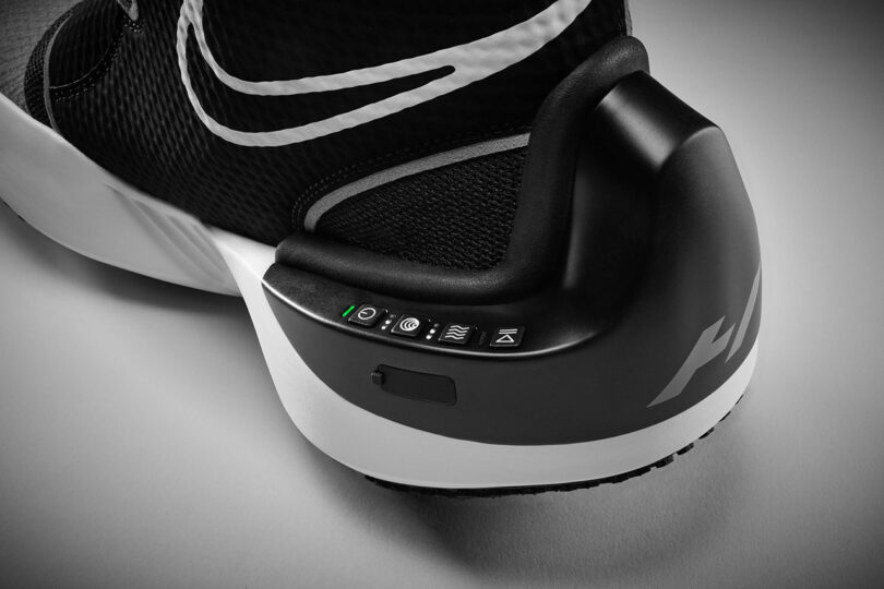 Close-up view of the heel area of a black and white Nike x Hyperice athletic shoe featuring electronic controls and an indicator light, showcasing advanced recovery technology.